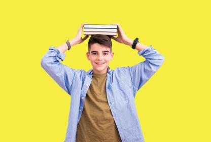 Usměvavý student drží na hlavě stoh knih, žluté pozadí