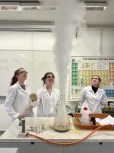 Studentky v laboratorních pláštích sledují pokus, ze kterého stoupá bílý dým